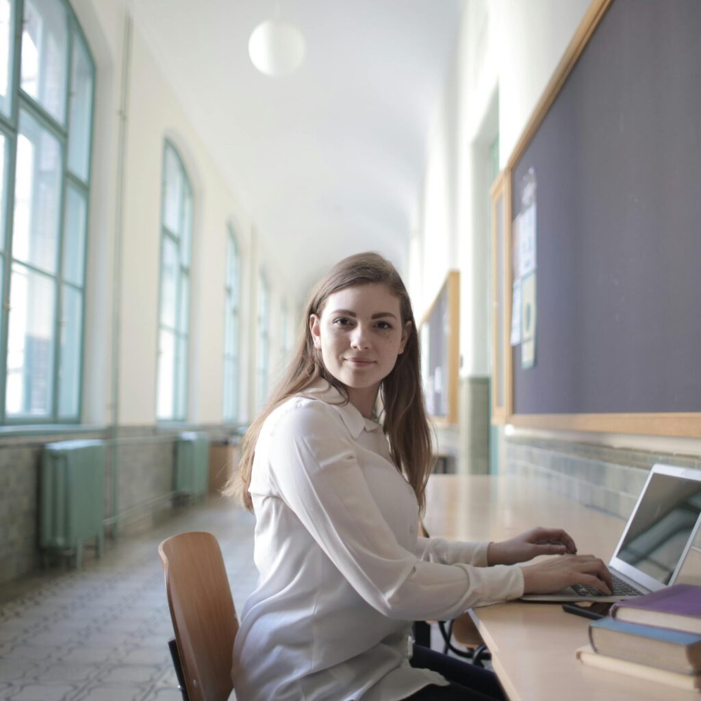 Side view of intelligent young woman in elegant wear sitting at table in university corridor and browsing netbook while working on assignment and looking at camera